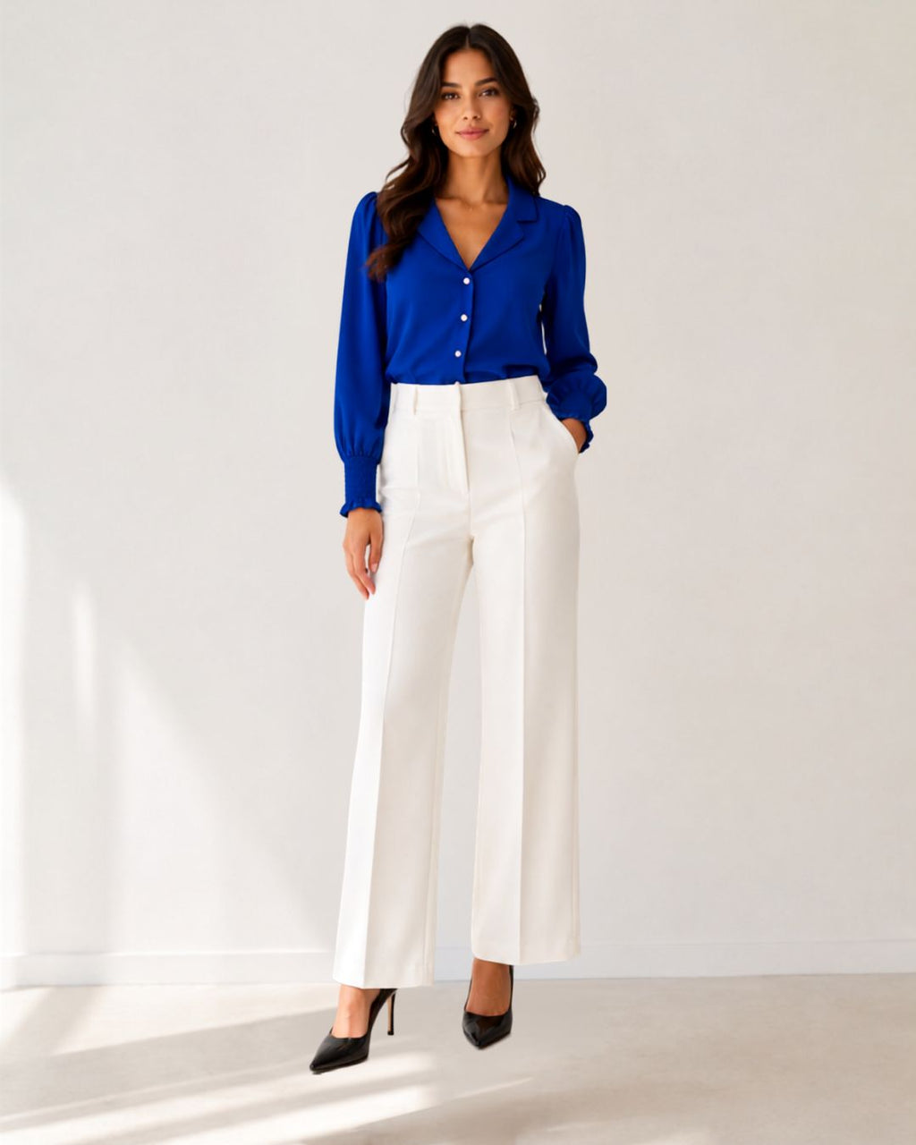 A woman modeling a vibrant royal blue long-sleeve button-up blouse paired with high-waisted white wide-leg trousers and black pointed-toe heels, standing against a clean white studio background.