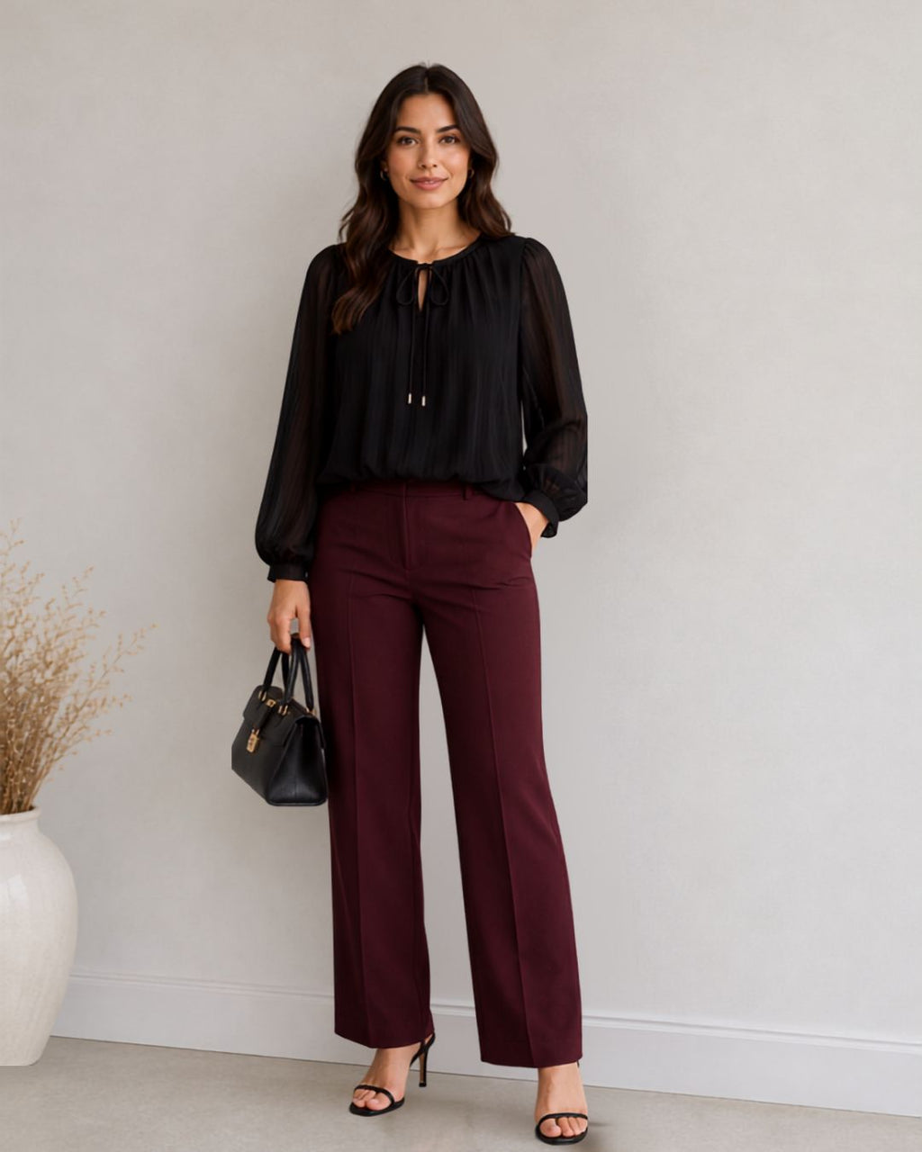 A woman stands against a light gray wall wearing a black sheer long-sleeve blouse and burgundy wide-leg trousers, holding a small black handbag.