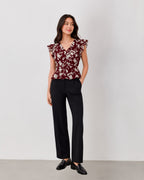 A woman modeling a burgundy floral short-sleeve top with ruffled shoulders and black wide-leg trousers, standing against a plain light gray studio background.