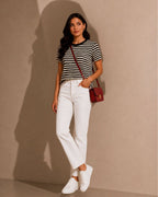 A woman stands against a beige wall wearing a black and white horizontally striped short-sleeve top, white straight-leg jeans, white sneakers, and carrying a small red crossbody bag.
