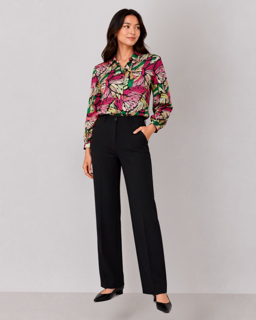 A woman modeling a long-sleeve button-up blouse with a vibrant pink, green, and yellow tropical floral print, paired with black wide-leg trousers and black pointed flats, against a light gray studio background.