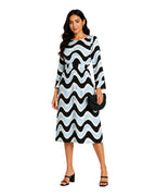 A woman modeling a knee-length dress with a bold black, white, and light blue wavy pattern, holding a black clutch purse and wearing black heeled sandals against a plain white background.