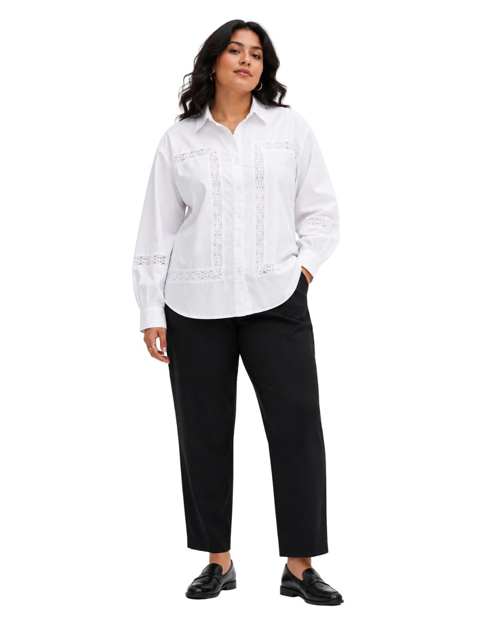 A woman modeling a white long-sleeve button-up shirt with vertical lace embroidery on the front, paired with black cropped pants and black loafers, against a plain white background.