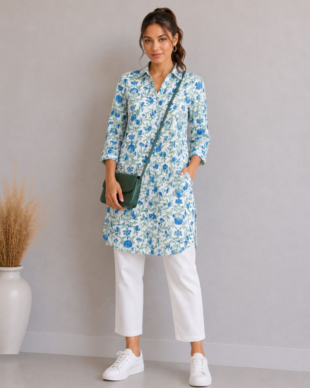 A woman models a light blue and white floral print tunic top with three-quarter sleeves, paired with white cropped pants, white sneakers, and a dark green crossbody bag, standing against a neutral gray wall.