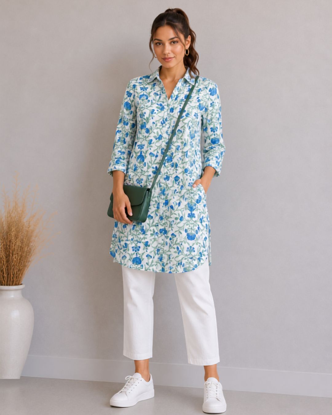 A woman models a light blue and white floral print tunic top with three-quarter sleeves, paired with white cropped pants, white sneakers, and a dark green crossbody bag, standing against a neutral gray wall.