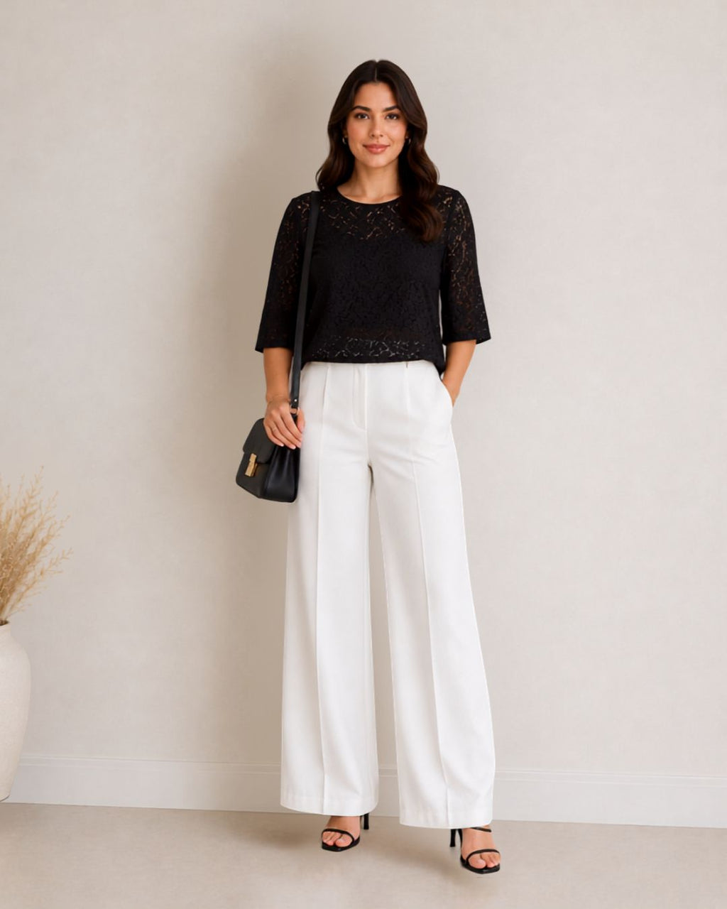 A woman stands against a plain light beige wall wearing a black lace three-quarter sleeve top and white wide-leg pants, holding a small black handbag.