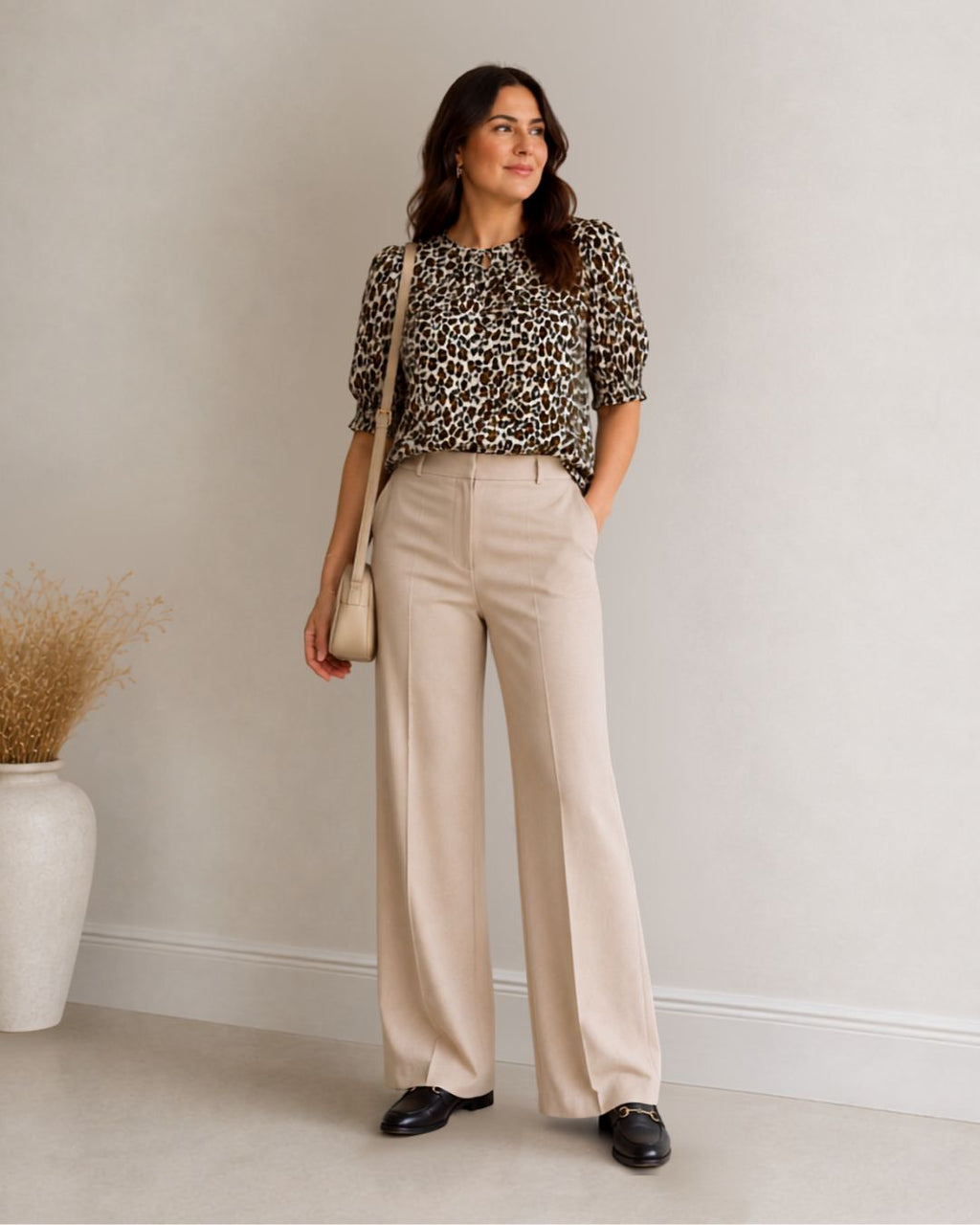 A woman stands in a studio wearing a leopard-print short-sleeve blouse, high-waisted beige wide-leg trousers, and black loafers, while holding a small beige shoulder bag.