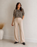A woman stands in a studio wearing a leopard-print short-sleeve blouse, high-waisted beige wide-leg trousers, and black loafers, while holding a small beige shoulder bag.