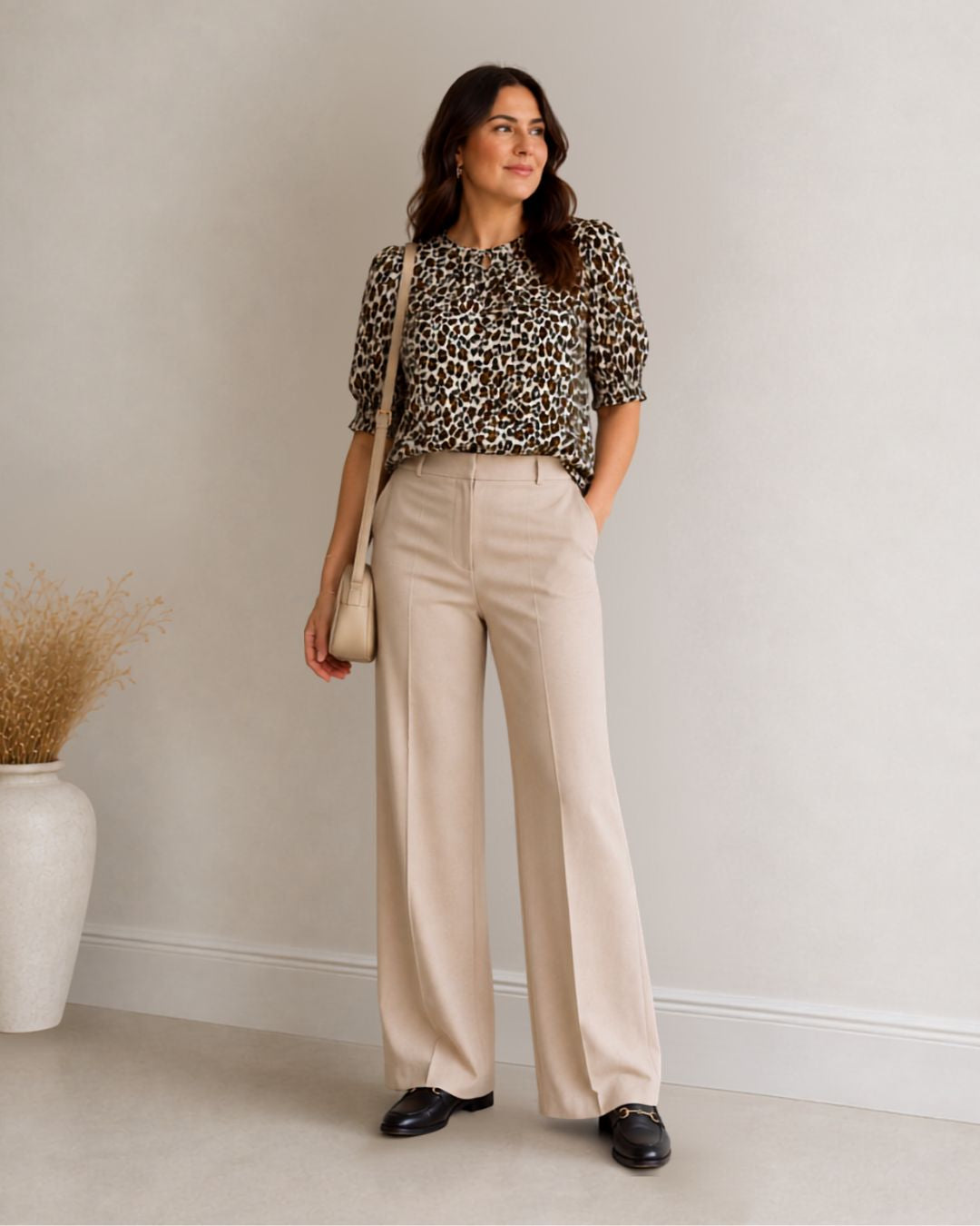 A woman stands in a studio wearing a leopard-print short-sleeve blouse, high-waisted beige wide-leg trousers, and black loafers, while holding a small beige shoulder bag.
