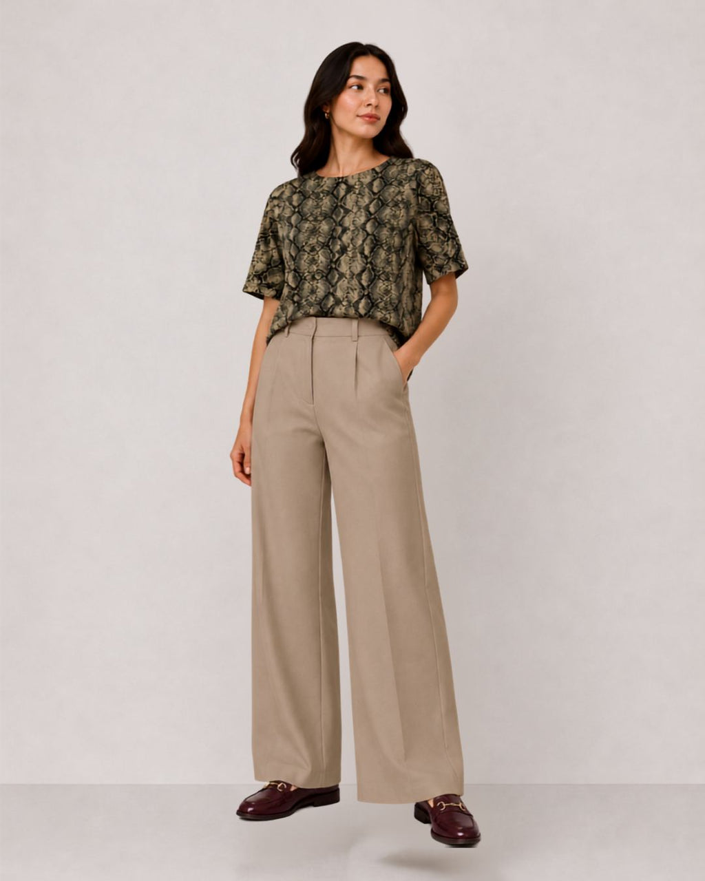 A woman modeling a short-sleeve top with an olive green and black snakeskin pattern, paired with high-waisted, wide-leg beige trousers and dark brown loafers, against a plain light gray studio background.