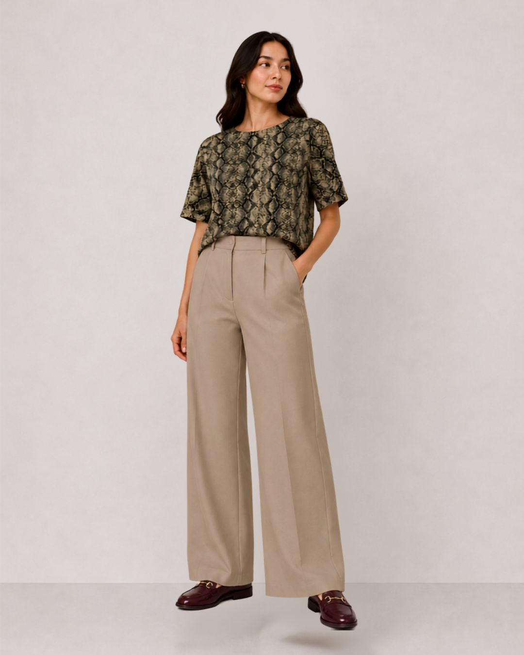 A woman modeling a short-sleeve top with an olive green and black snakeskin pattern, paired with high-waisted, wide-leg beige trousers and dark brown loafers, against a plain light gray studio background.