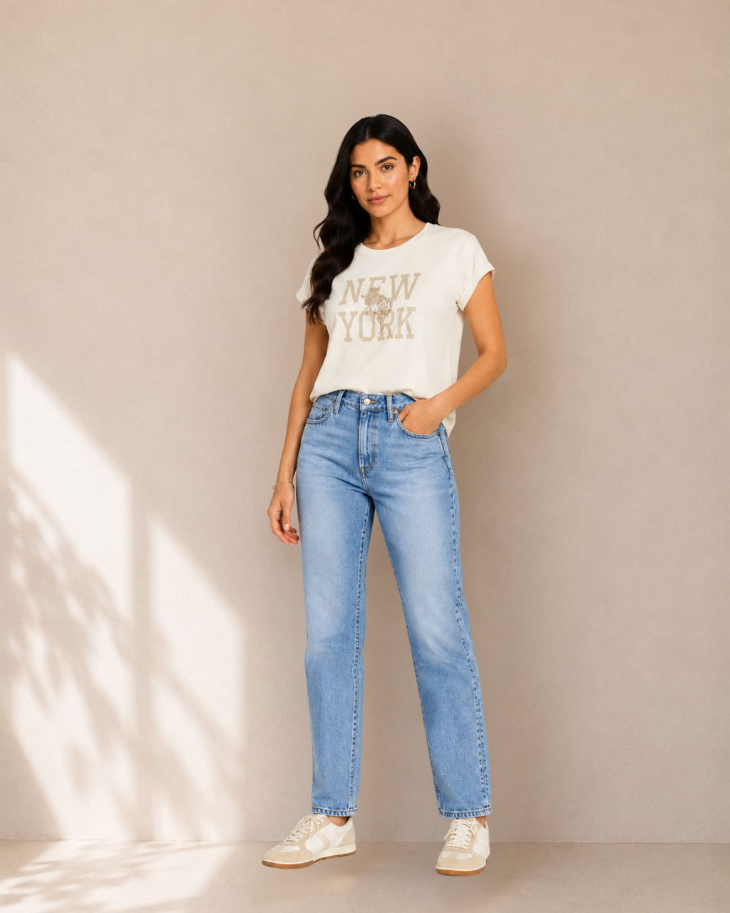 A woman modeling a white graphic t-shirt with 'NEW YORK' printed in gold, paired with light-wash blue jeans and white sneakers, standing against a beige wall with soft shadow projections.