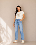A woman modeling a white graphic t-shirt with 'NEW YORK' printed in gold, paired with light-wash blue jeans and white sneakers, standing against a beige wall with soft shadow projections.