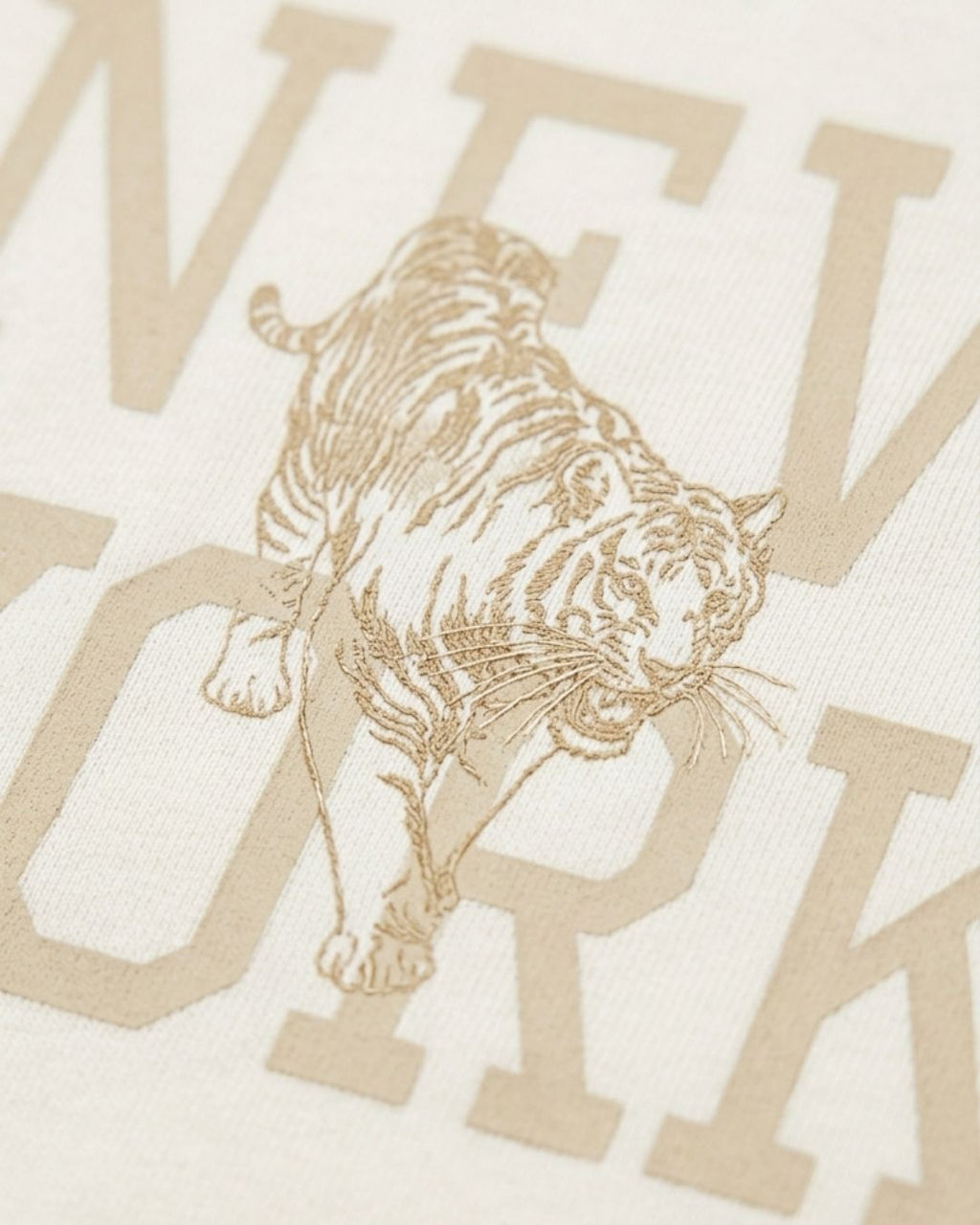 Close-up of a cream-colored fabric with a tan/golden graphic print featuring a walking tiger and large partial letters spelling a city name.