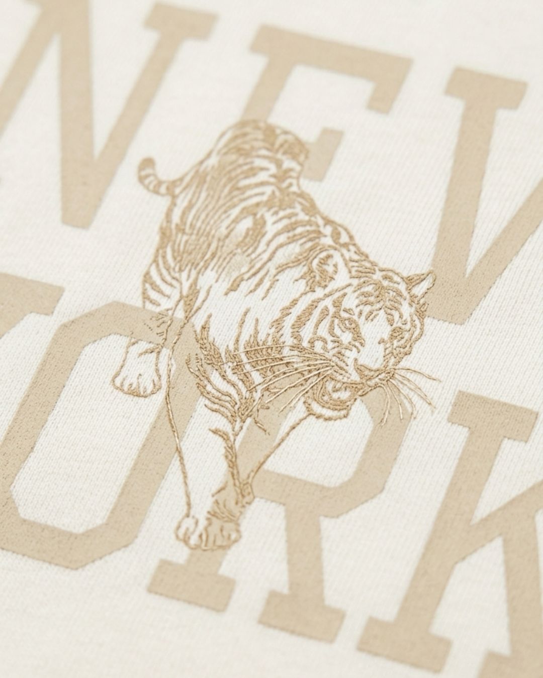 Close-up of a cream-colored fabric with a tan/golden graphic print featuring a walking tiger and large partial letters spelling a city name.