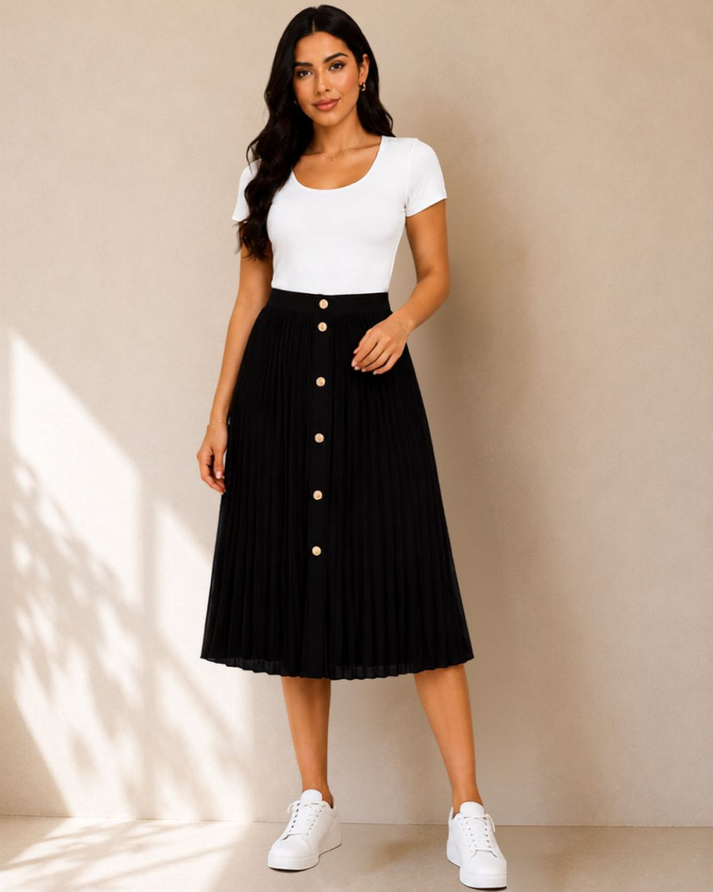 A woman modeling a white short-sleeve scoop-neck top paired with a black pleated midi skirt featuring a row of gold buttons down the front, standing against a beige wall with soft window shadows.