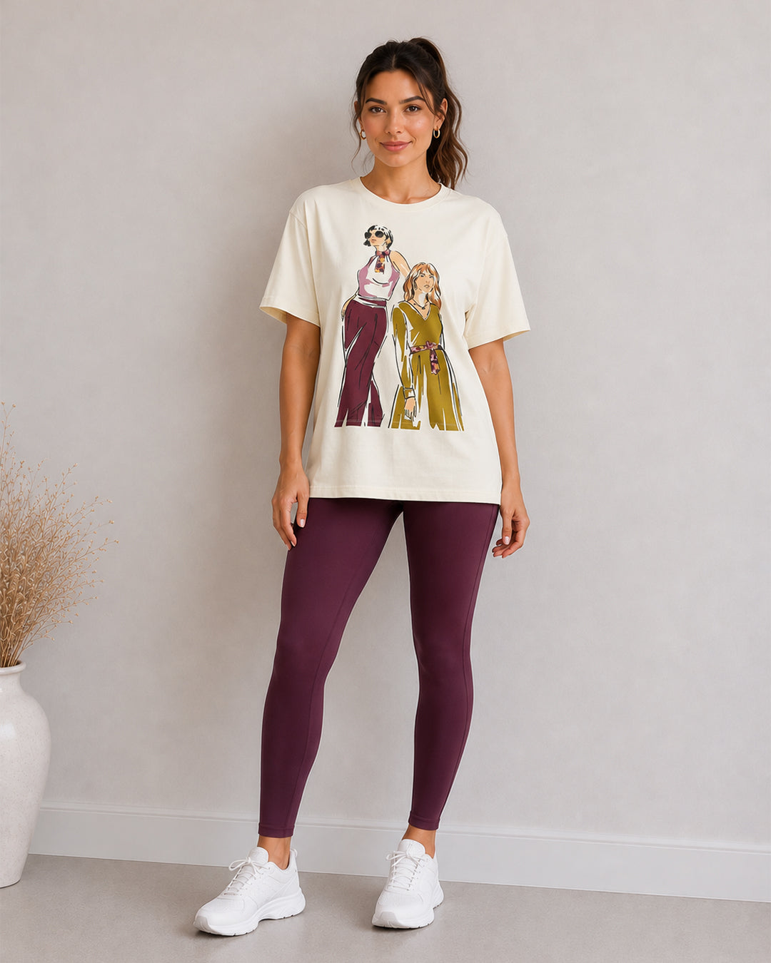 A woman modeling a cream graphic t-shirt with a retro illustration of two women, paired with dark purple leggings and white sneakers, standing against a light gray wall.