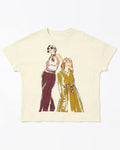 An off-white short-sleeve t-shirt featuring a graphic print of two retro-style illustrated women, one in a pink and maroon outfit and the other in a yellow-green dress, both wearing patterned neckties.
