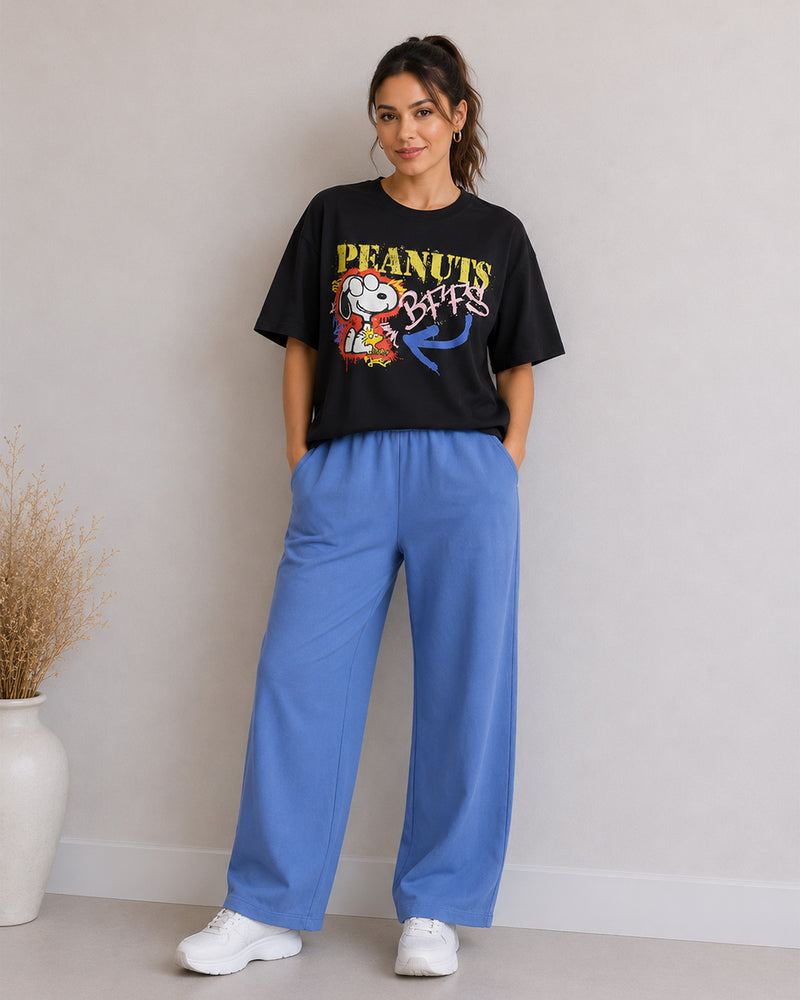A woman modeling a black graphic t-shirt with a colorful 'Peanuts' print and wide-leg blue pants, standing in a studio setting.