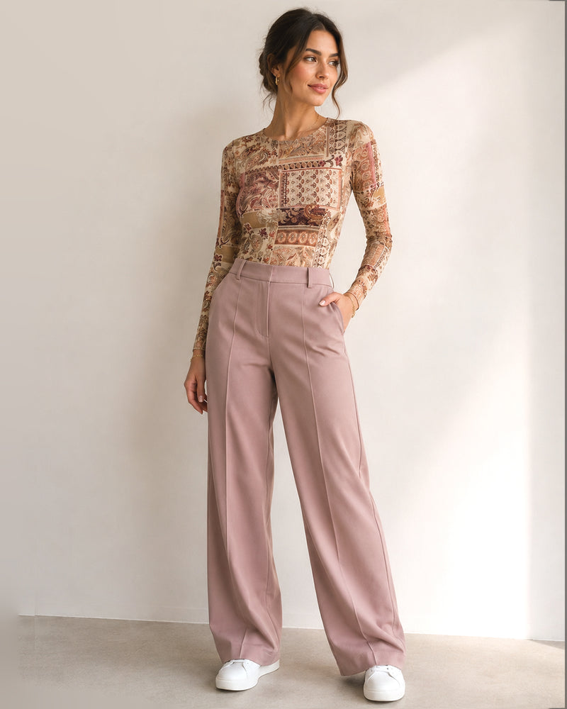 A woman modeling a long-sleeve beige and brown patterned top paired with dusty pink wide-leg trousers and white sneakers, standing against a plain off-white wall.