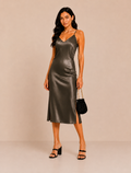 A woman modeling a dark olive green satin slip dress with spaghetti straps, holding a small black beaded handbag and wearing black strappy heels against a solid beige studio background.