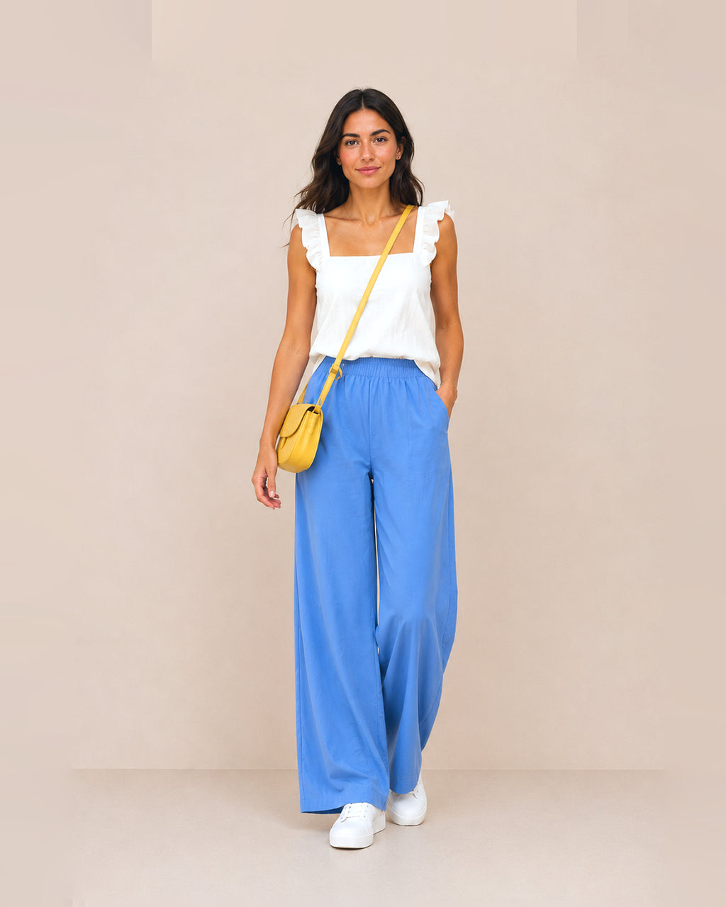 A woman stands against a plain beige studio background wearing a white ruffled-strap sleeveless top, bright blue wide-leg pants, white sneakers, and a small yellow crossbody bag.