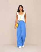 A woman stands against a plain beige studio background wearing a white ruffled-strap sleeveless top, bright blue wide-leg pants, white sneakers, and a small yellow crossbody bag.