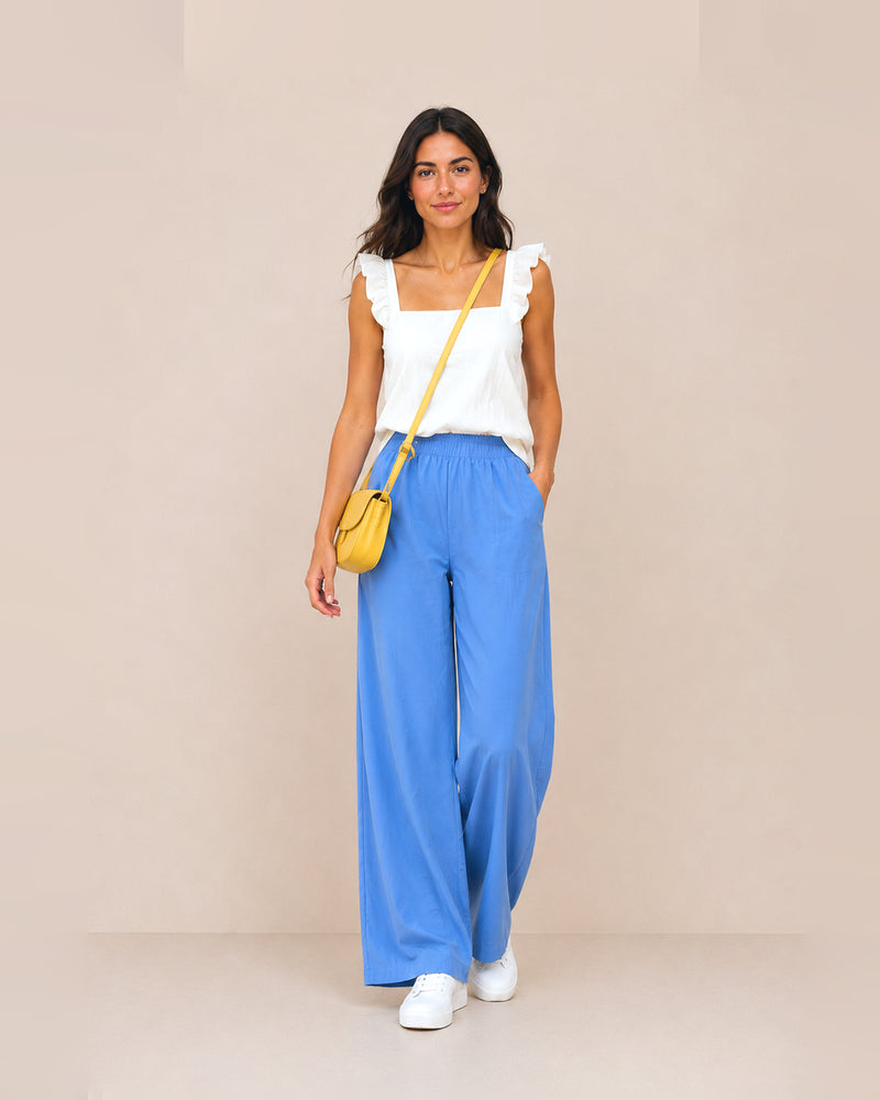 A woman stands against a plain beige studio background wearing a white ruffled-strap sleeveless top, bright blue wide-leg pants, white sneakers, and a small yellow crossbody bag.