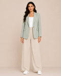 A woman modeling a sage green blazer, cream wide-leg trousers, a white top, and white sneakers against a solid beige studio background.