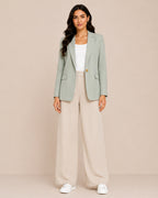 A woman modeling a sage green blazer, cream wide-leg trousers, a white top, and white sneakers against a solid beige studio background.