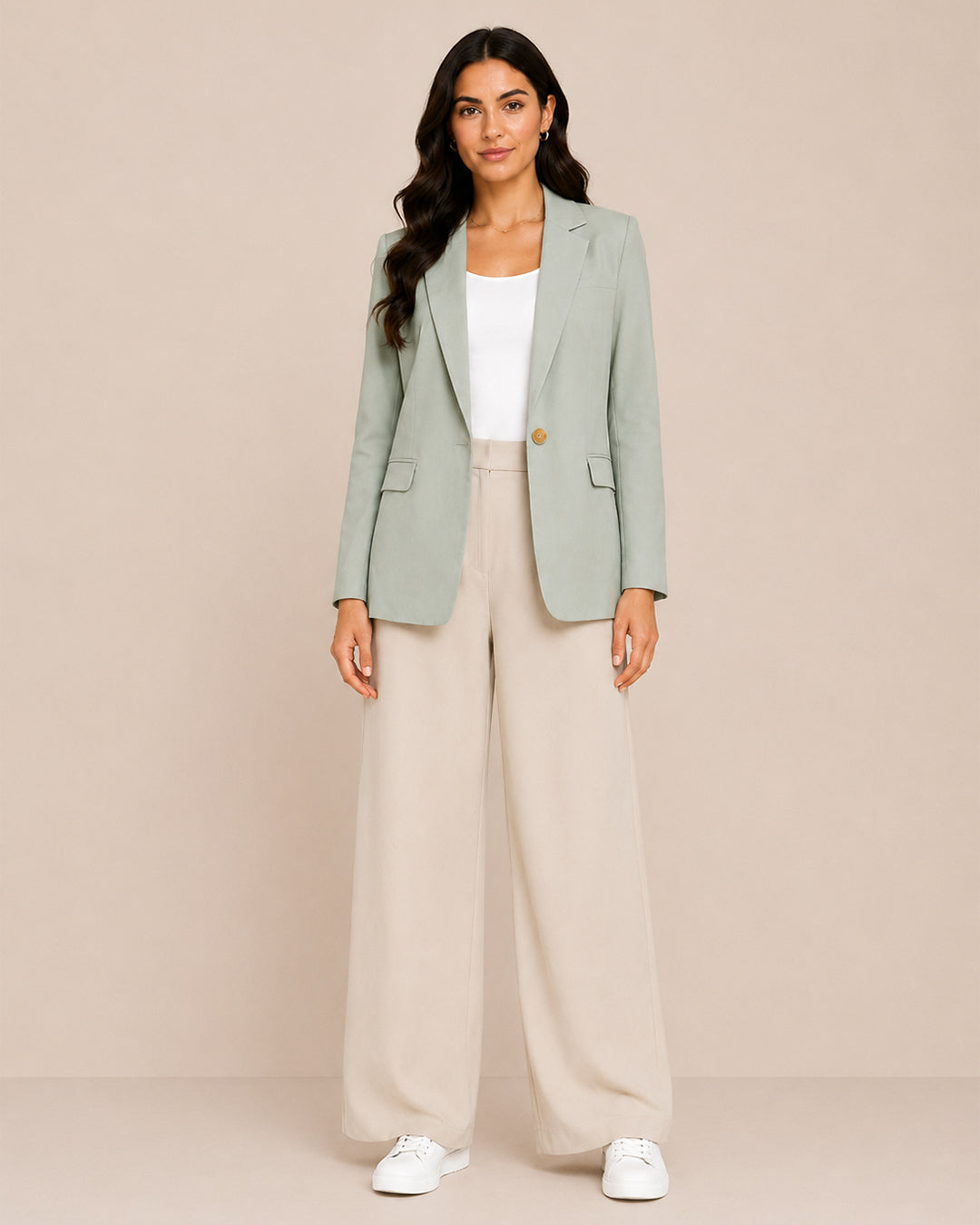 A woman modeling a sage green blazer, cream wide-leg trousers, a white top, and white sneakers against a solid beige studio background.