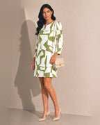 A woman models a long-sleeve mini dress with an abstract geometric pattern in sage green and white. She is holding a small beige handbag and wearing beige high heels, standing against a plain beige wall with a soft shadow.