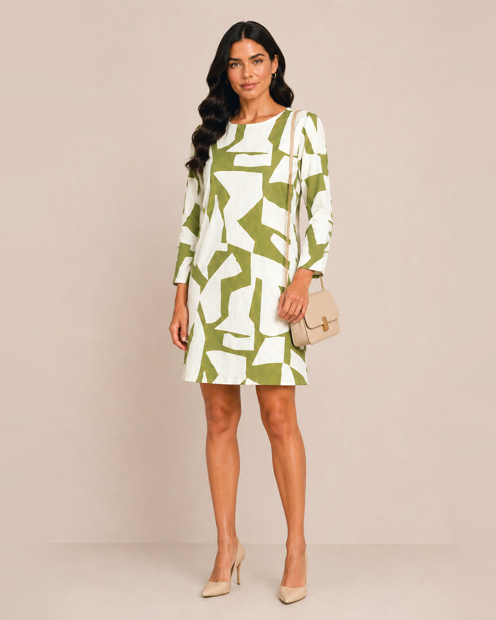 A woman modeling a knee-length shift dress with a bold abstract green and white geometric pattern, holding a small beige handbag and wearing beige heels against a solid beige studio background.
