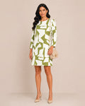 A woman modeling a knee-length shift dress with a bold abstract green and white geometric pattern, holding a small beige handbag and wearing beige heels against a solid beige studio background.