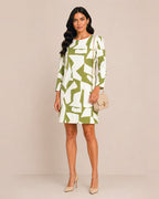 A woman modeling a knee-length shift dress with a bold abstract green and white geometric pattern, holding a small beige handbag and wearing beige heels against a solid beige studio background.