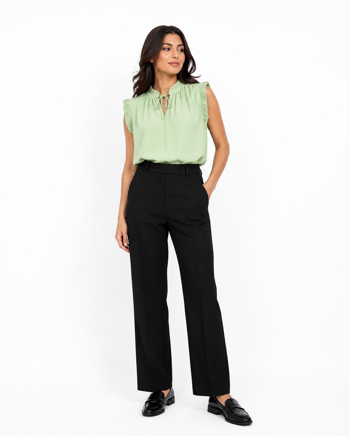 A woman modeling a light green sleeveless blouse with a tie-neck detail and black wide-leg trousers, standing against a plain white studio background.