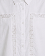Close-up of a white button-front shirt with decorative lace trim running vertically on the chest and across the pocket edges.
