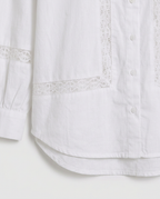 Close-up of a white button-front shirt with delicate lace trim along the pocket edges and sleeve cuff, shown against a plain light background.