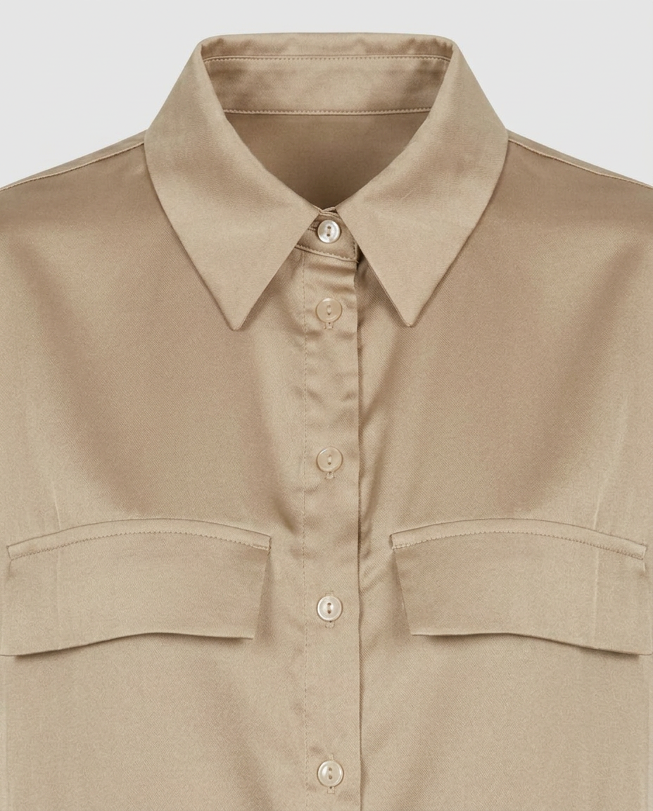 A close-up of a beige button-up shirt with a pointed collar and two chest flap pockets, shown against a light gray background.