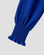 A close-up of a royal blue garment sleeve with a gathered smocked cuff and ruffled hem, shown against a light gray background.