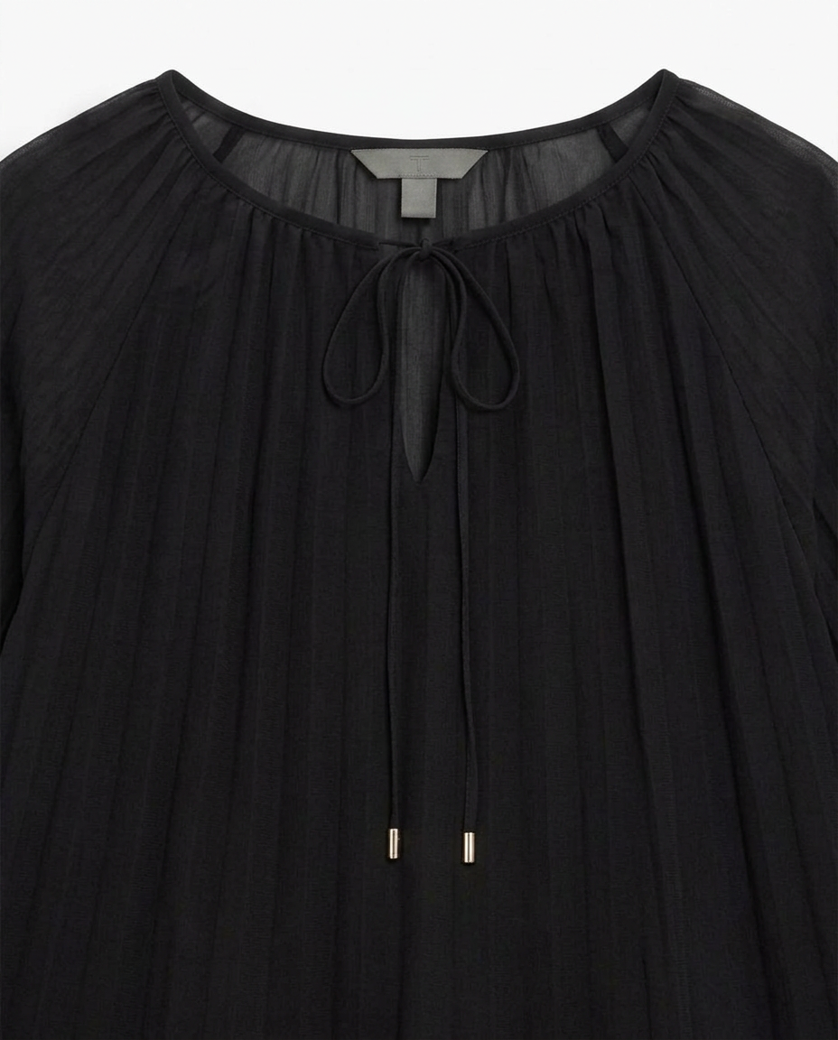 A close-up of a black pleated blouse with a gathered round neckline, a front tie closure, and small metallic gold ring accents near the center front.