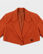 A cropped orange-brown blazer with notched lapels and a single visible dark button, laid flat against a light gray background.