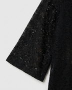 Close-up of a black lace garment sleeve with sheer floral embroidery and subtle shimmer, shown against a light gray background.